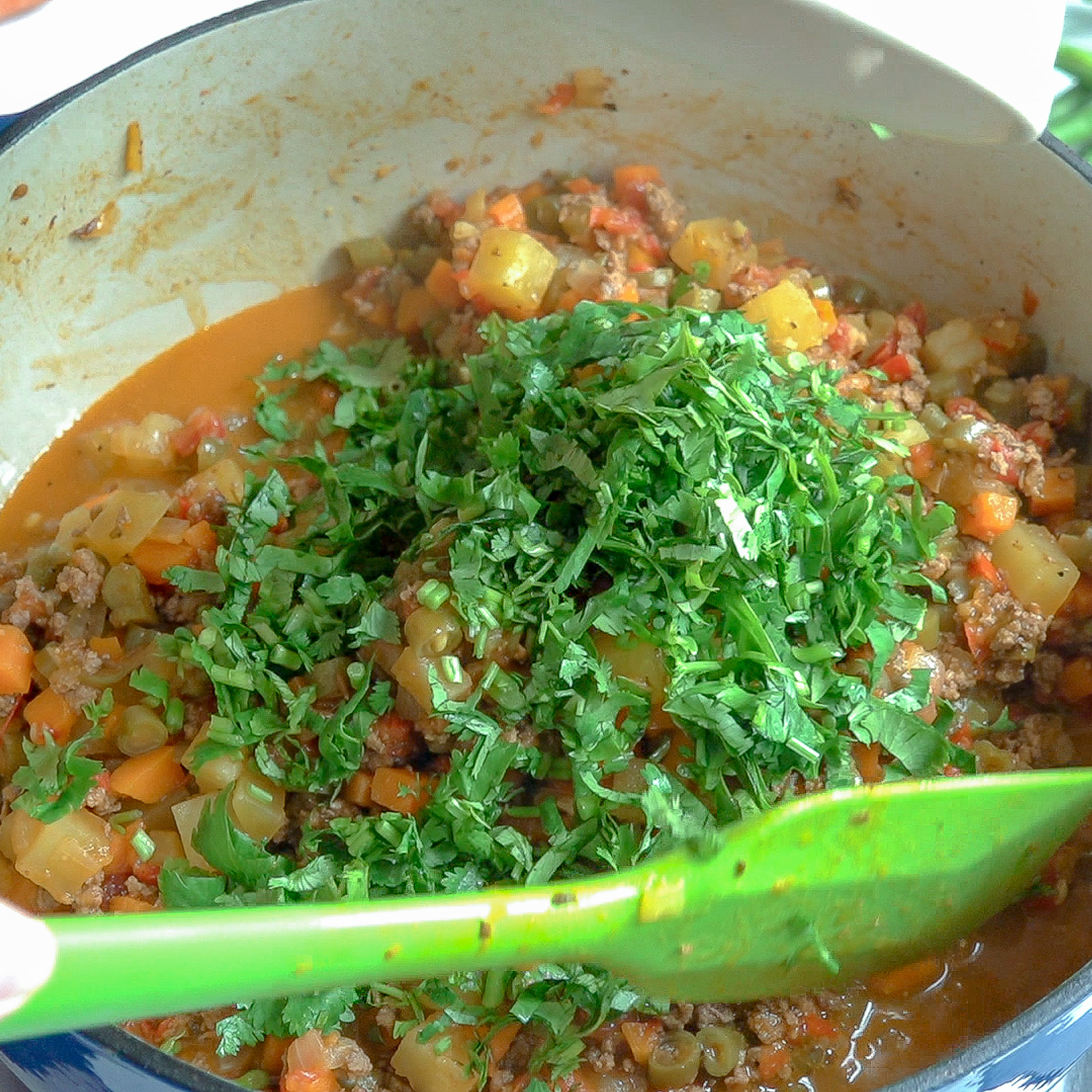 Olla grande presentando el picadillo cocinado, arriba un puño de cilantro y una espatula verde que revuelve los ingredientes