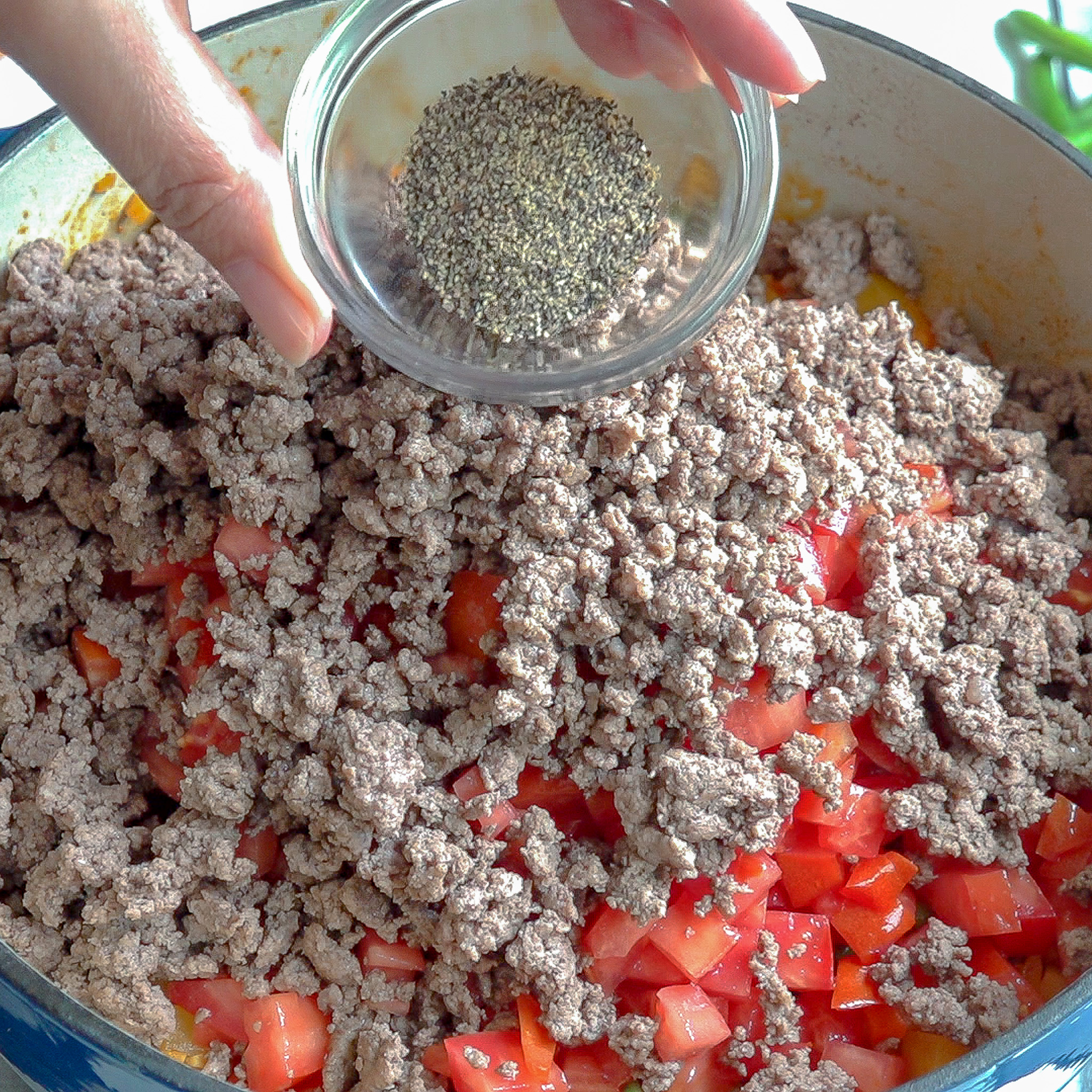 Una olla grande con todos los ingredientes, pero arriba sobresalen los tomates, la carme molida cocinada y una mano agrega semillas de comino