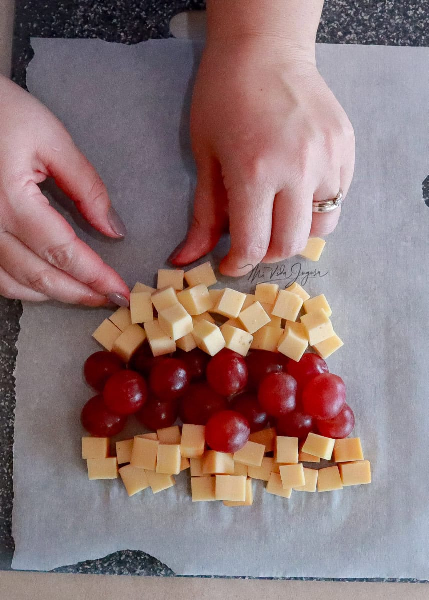 Tres lineas de ingredientes, la mas grande quesos, la del medio uvas y la mas pequeña otro tipo de queso, comenzando a formar un triangulo para simular la forma de arbol