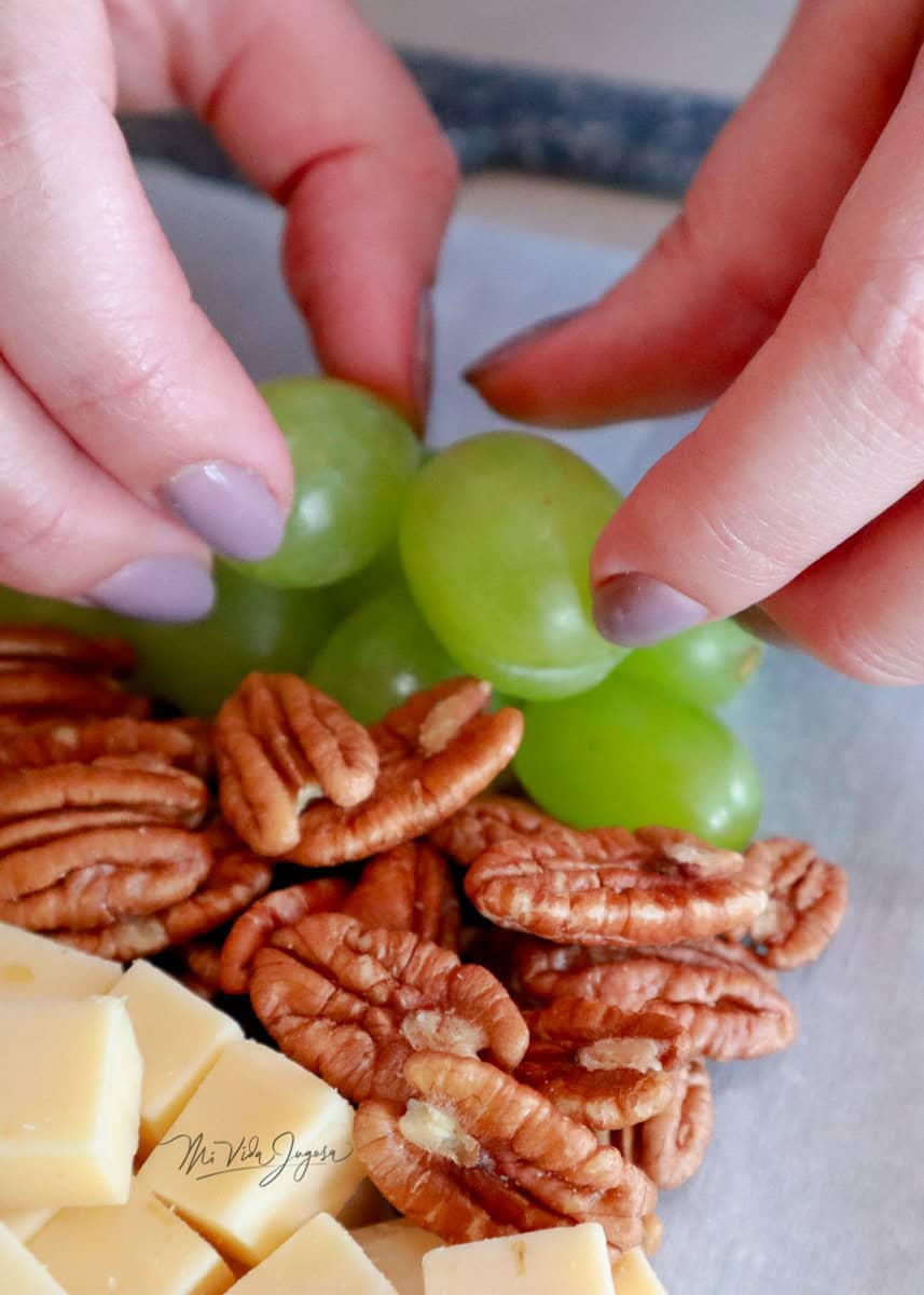 Dos manos acomodando en linea uvas verdes, estan colocadas a la par de pecanas formadas en linea.