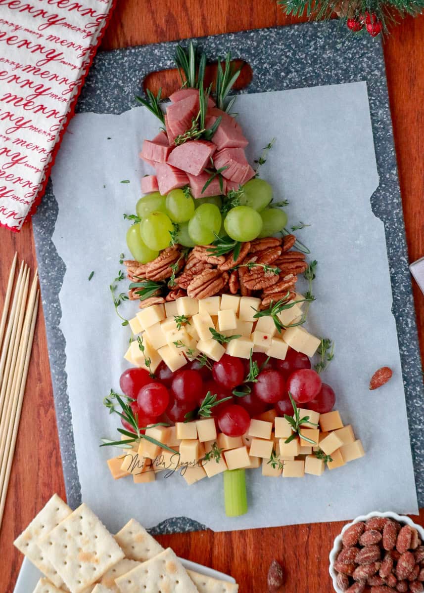 Quesos, frutas, semillas y embutidos colocados en forma de arbol de navidad, sobre una tabla negra y papel encerado, al lado acompañado de galletas saladas y una taza de nueces