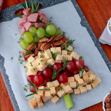 Una tabla de quesos con embutidos y frutas frescas con semillas decoradas en forma de arbol de navidad, un palo de apio simulando el palo del arbol de navidad