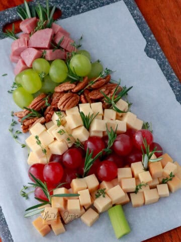 Una tabla de quesos con embutidos y frutas frescas con semillas decoradas en forma de arbol de navidad, un palo de apio simulando el palo del arbol de navidad