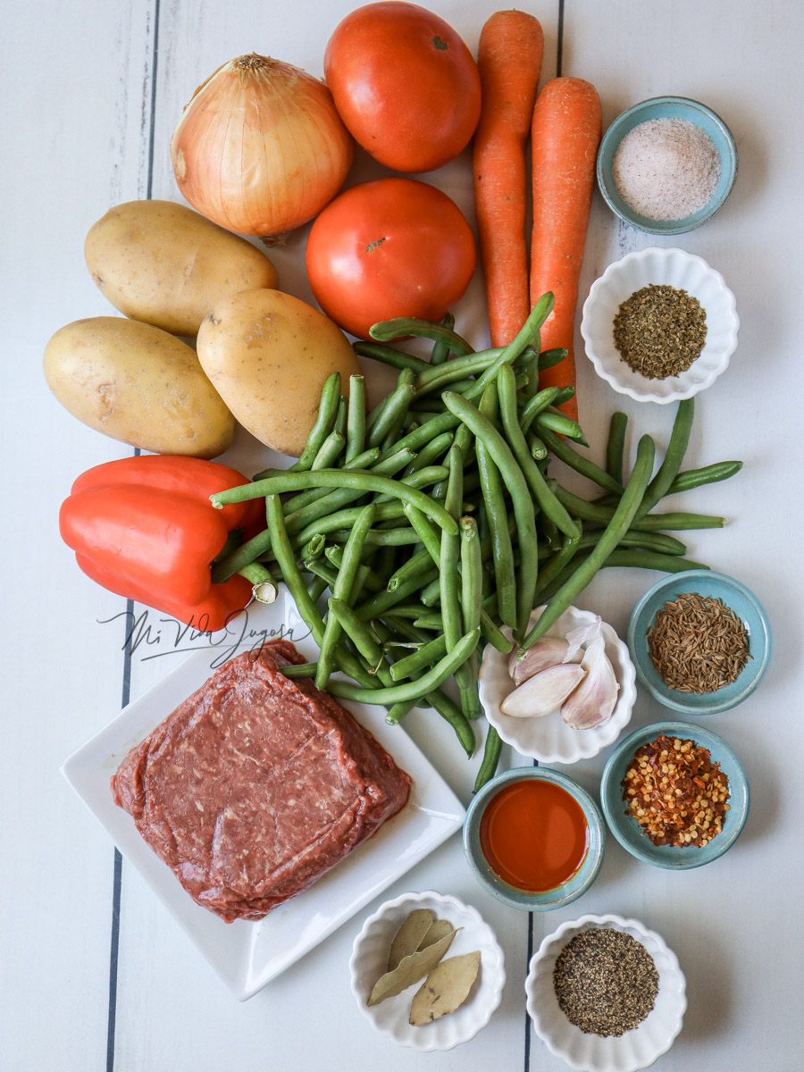 Todos los ingredientes se van a utilizar en la receta: Vainicas, carne de res, zanahoria, tomates, papas, etc. Estan colocados sobre una mesa blanca