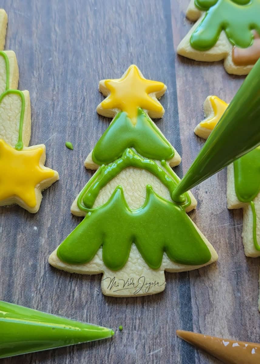 Galleta de mantequilla en forma de arbolito de navidad con estrella amarilla. Una manga pastelera rellena de glaseado real decora el arbol de color verde