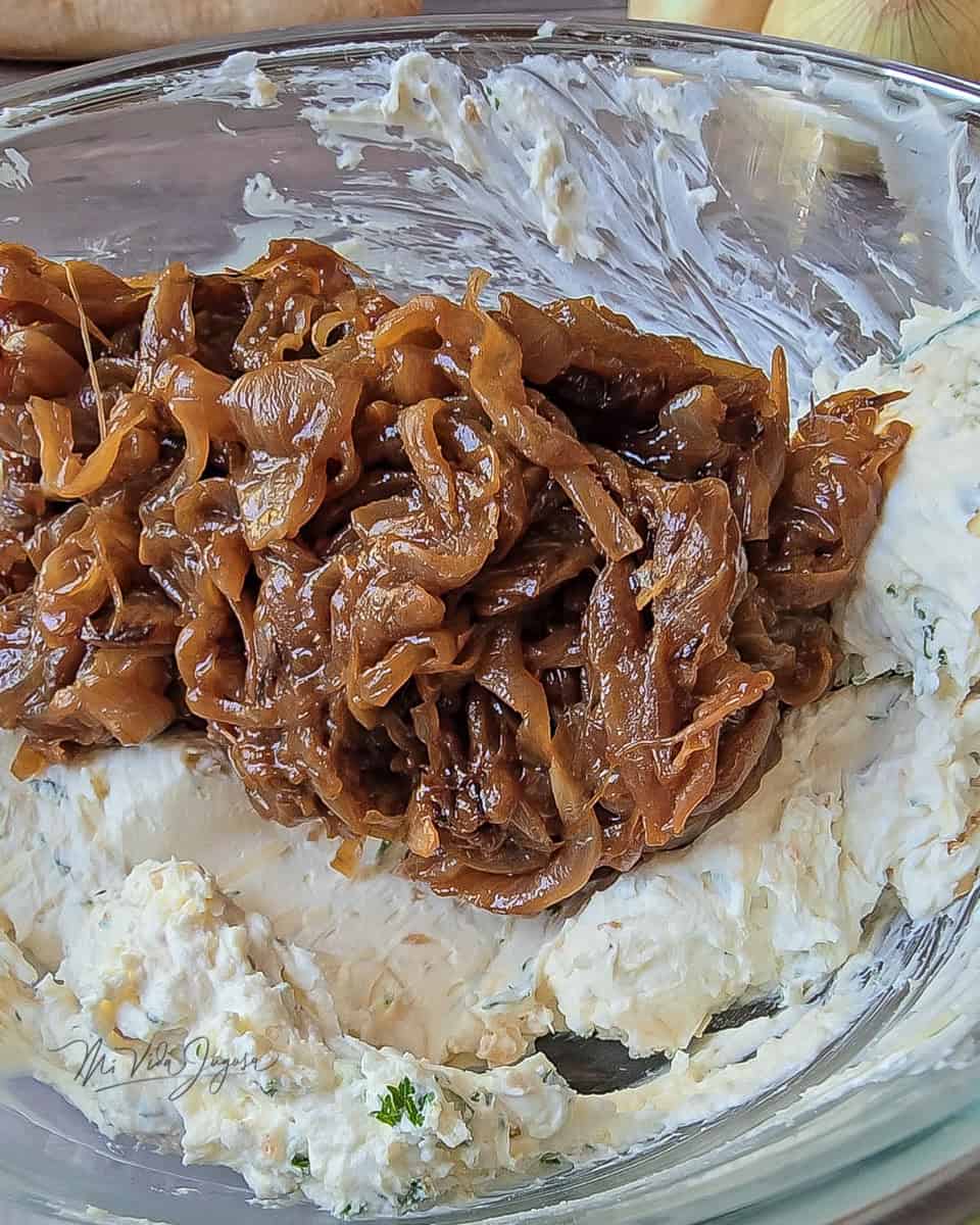 Cebolla caramelizada recién cocinada sobre una mezcla de queso crema con perejil, lista para ser combinada en un tazon de vidrio.