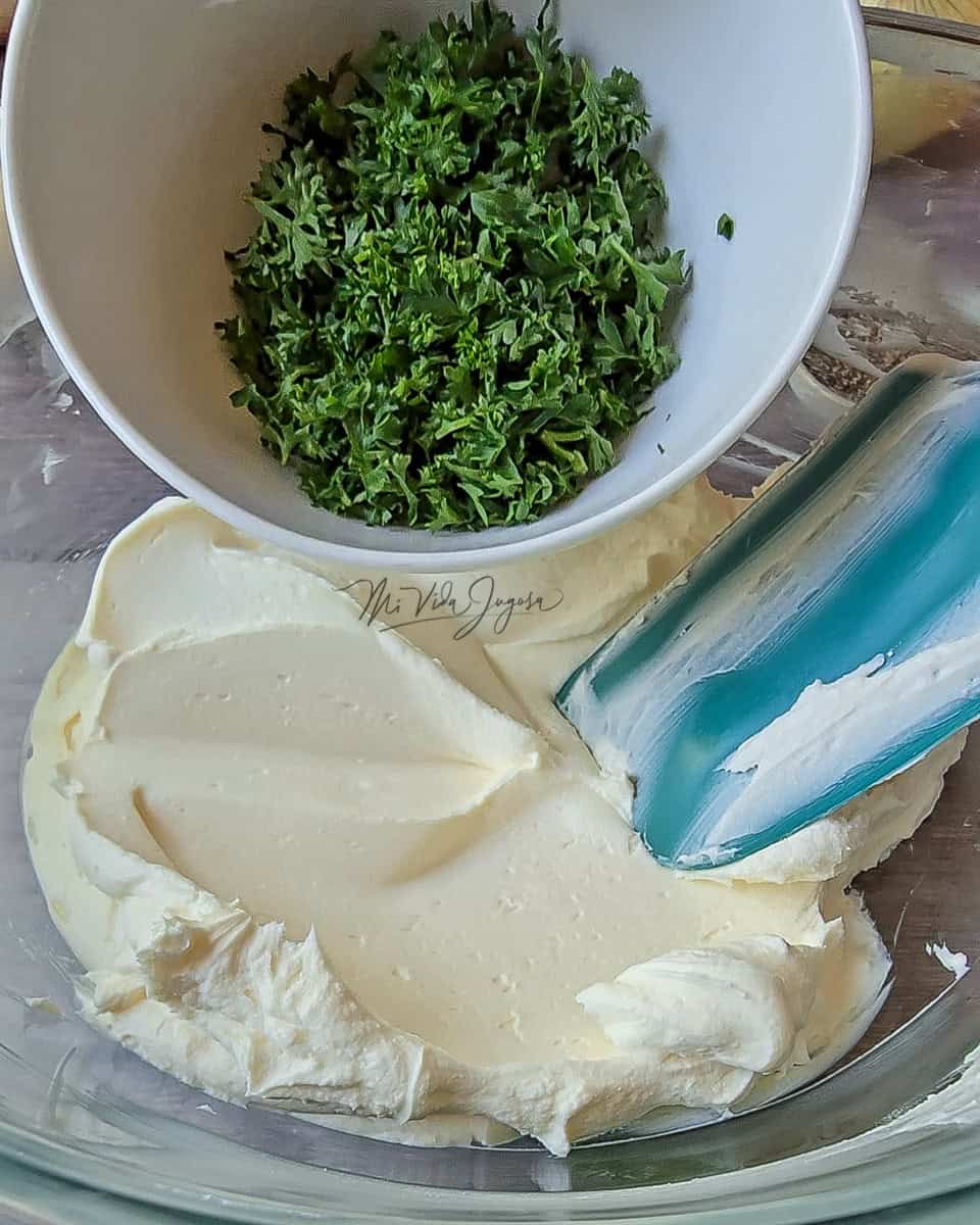 En un tazón transparente que contiene queso crema a temperatura ambiente, se esta agregando el perejil finamente picado