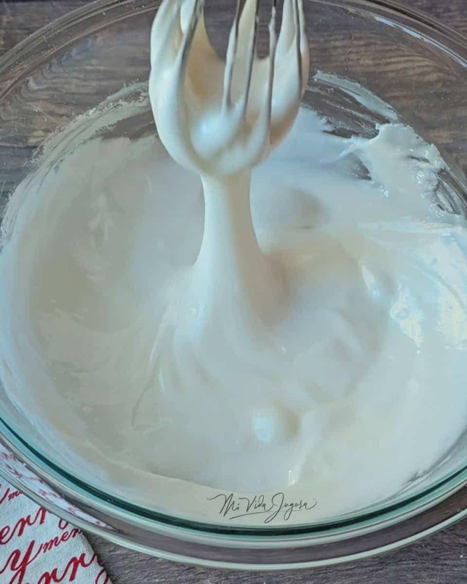 Un tazon transparente con glaseado real sin claras de huevo de color blanco. Además, una espatula de globo alzada mostrando la consistencia del glaseado