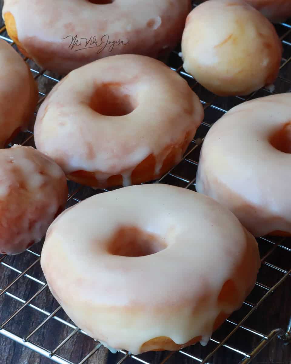 Donas cocidas y listas para degustar, decoradas con glas en la parte superior, presentadas sobre una bandeja cuadriculada, que permite el enfriado.

