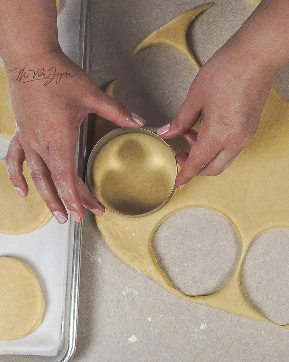 Masa extendida sobre una superficie plana, y dos manos que sostienen un cortado de forma circular que corta la masa aplanada previamente