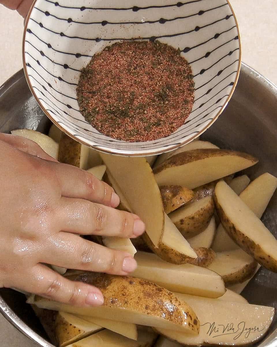 Un tazon lleno de papas cortadas en gajo, y una tacita con condimentos, listos para ser agregados al tazon.