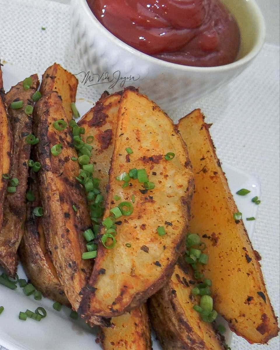 Papas cortadas en forma de gajo, cocinas y servidas sobre un plato blanco, decoradas con cebollinos finamente cortados, y una taza al lado conteniendo salsa de tomate tipo ketchup.