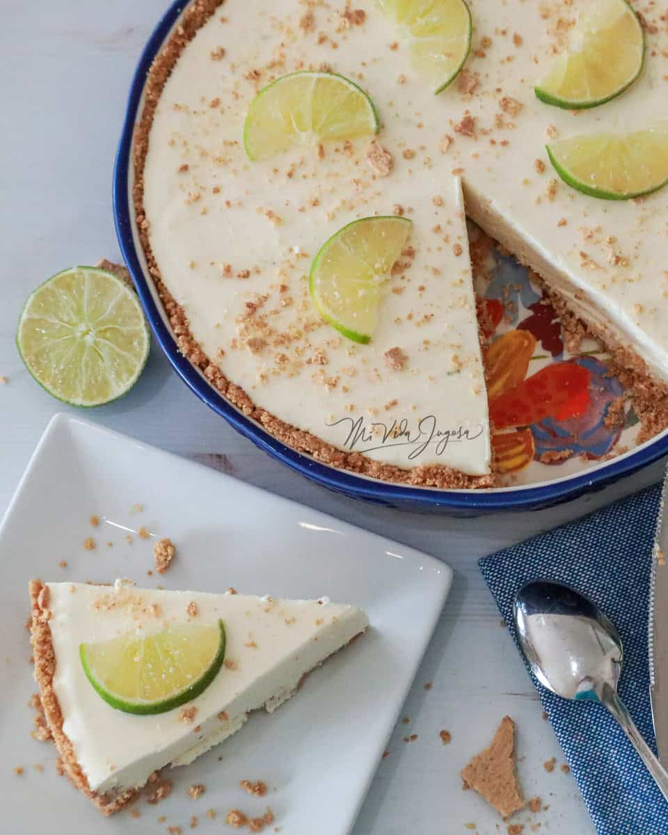 Pay de limón decorado con medias lunas de limón y boronas de galleta triturada, al frente un pedazo del pay servido en un plato blanco