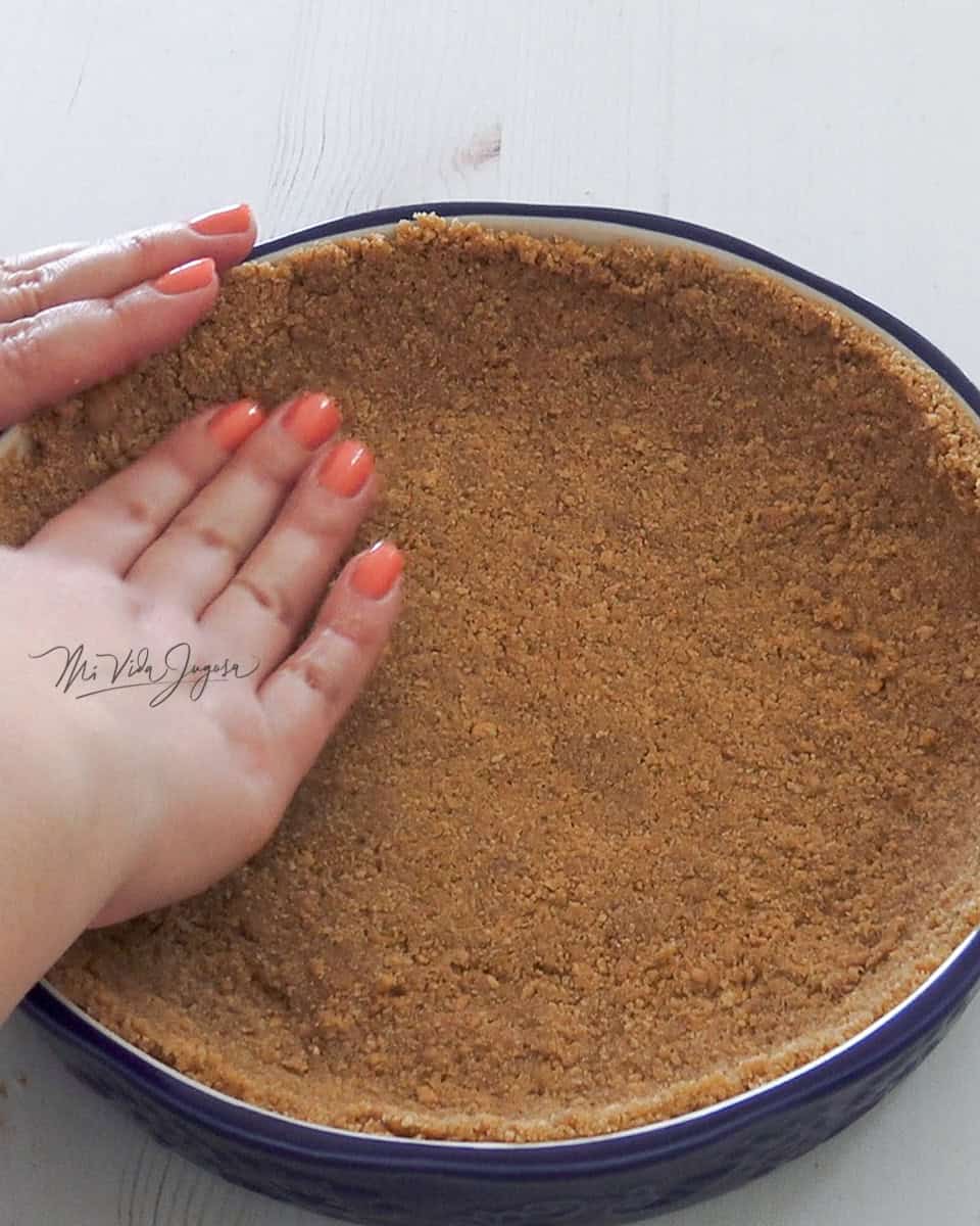 Mezcla de galleta siendo extendida con las manos por todo el fondo de un molde para pay color azul