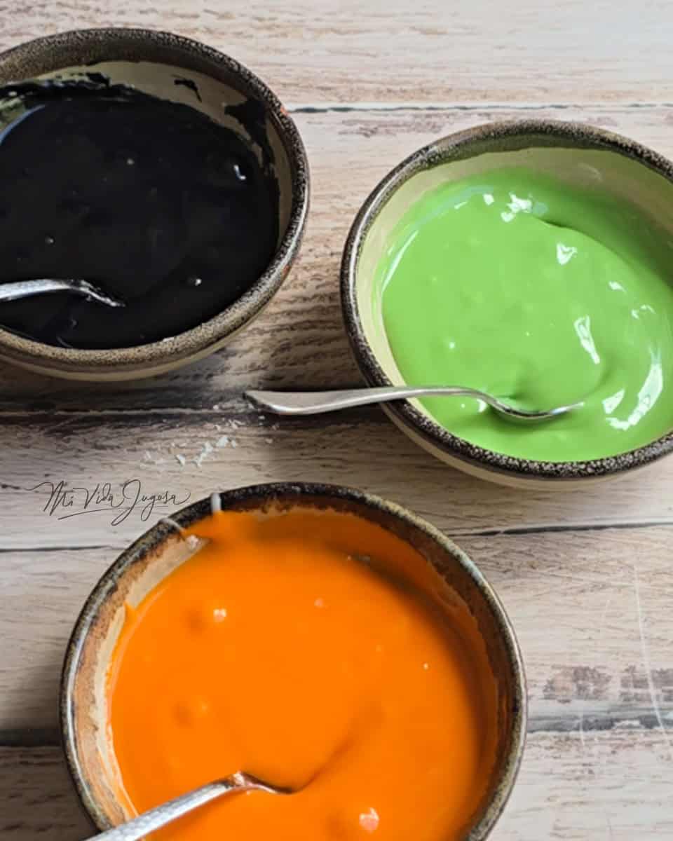 Tres tacitas de vidrio las cuales contienen tres colores de glaseado, siendo negro, verde y naranja
