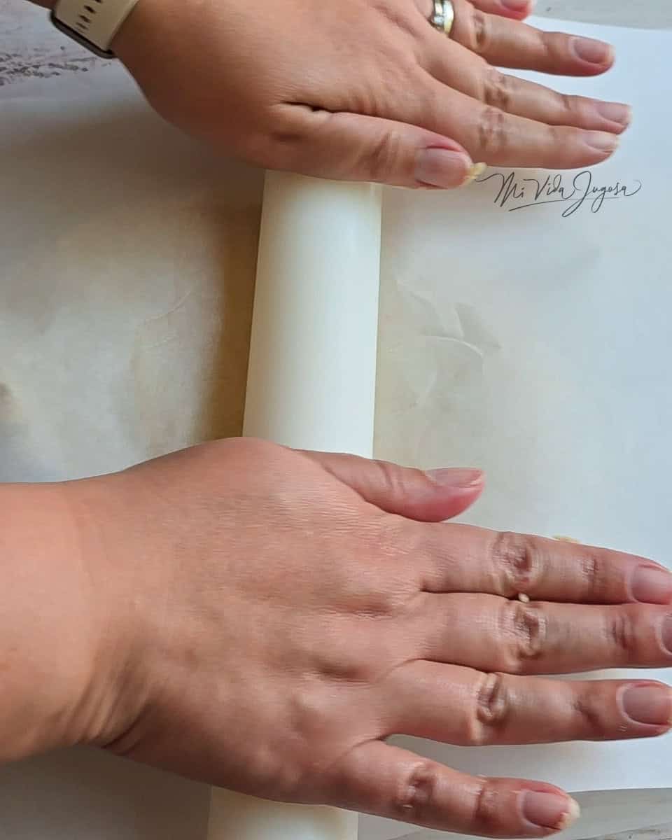 Se observan las manos sosteniendo un rodillo, el cual se encuentra estirando la masa que se encuentra en medio de los pliegos de papel encerado 