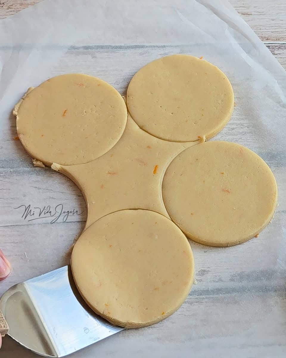 Masa aplanada, cortada en forma circular, en donde se observa una espatula levantando la forma de galleta y separandola del resto de la masa