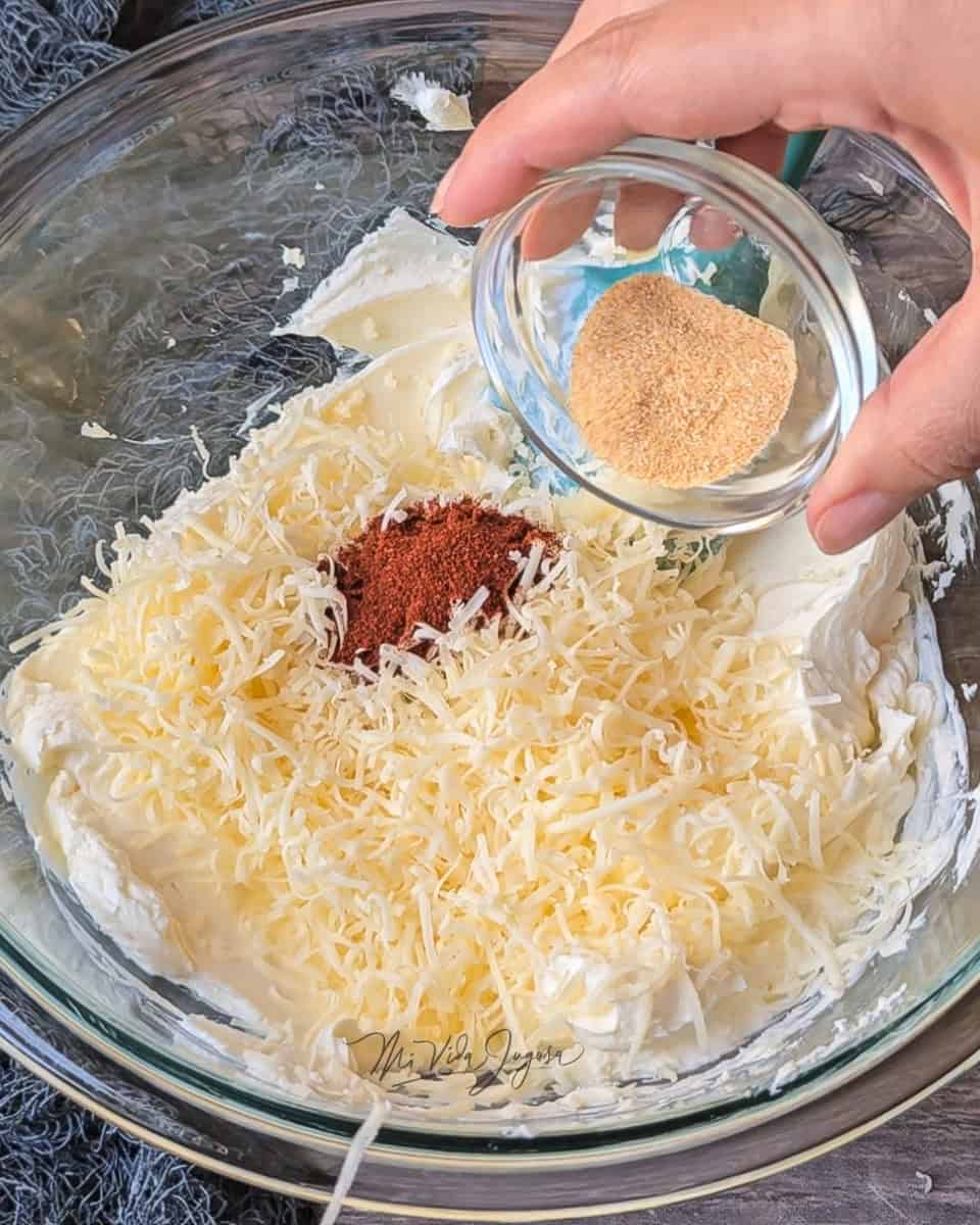 El bowl de vidrio con queso crema, donde se le estan agregando el queso cheddar, el chile chipotle, el ajo y los demas condimentos.