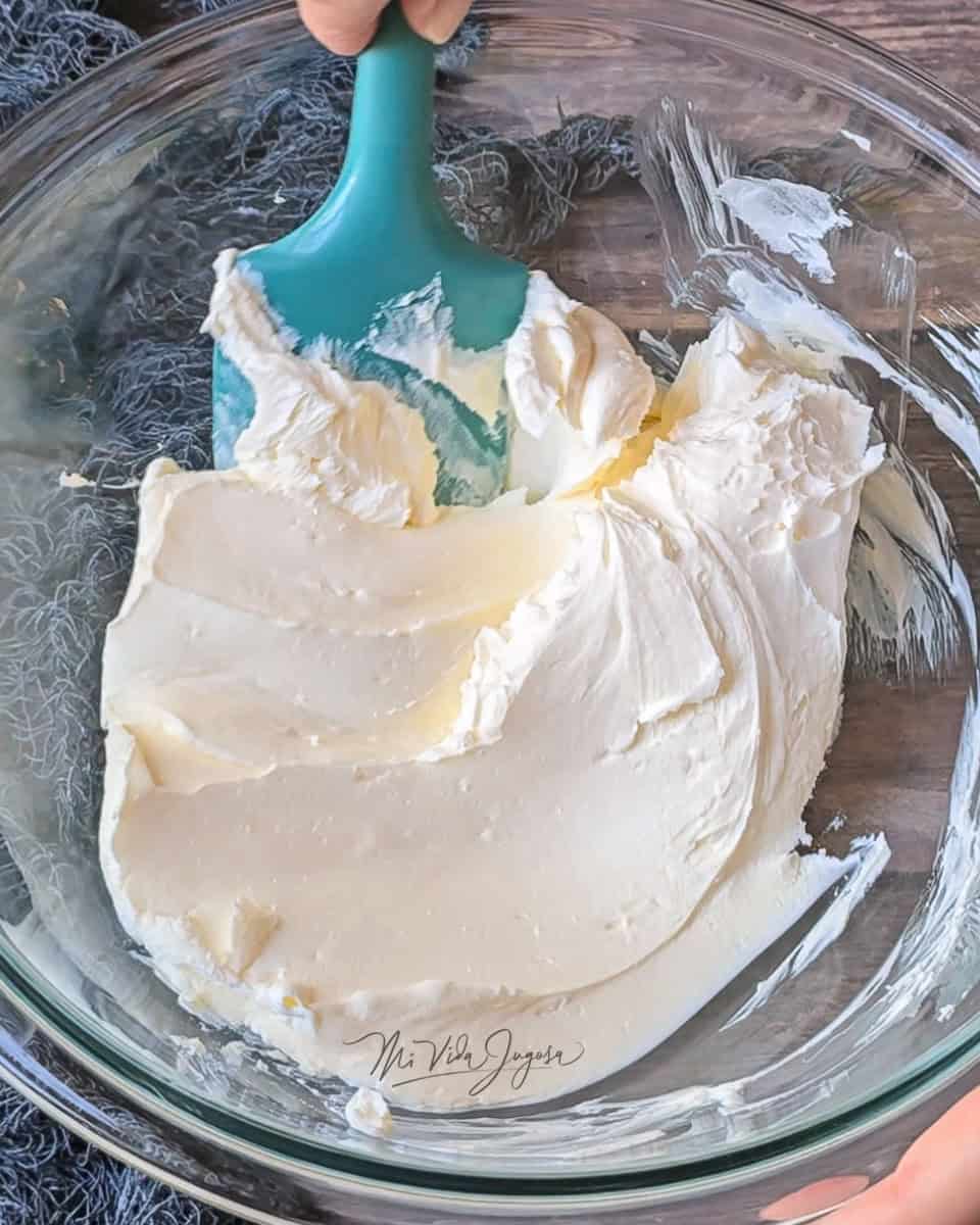Un bowl de vidrio con queso crema y una espatula que muestra estarlo batiendo