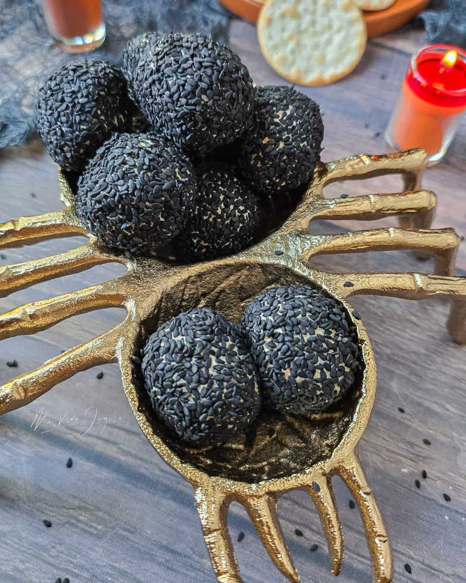 Se muestra las bolas de queso crema con queso cheddar y demas ingredientes, envueltas en semillas de ajonjolí negras, colocadas sobre una figura de araña decorativa