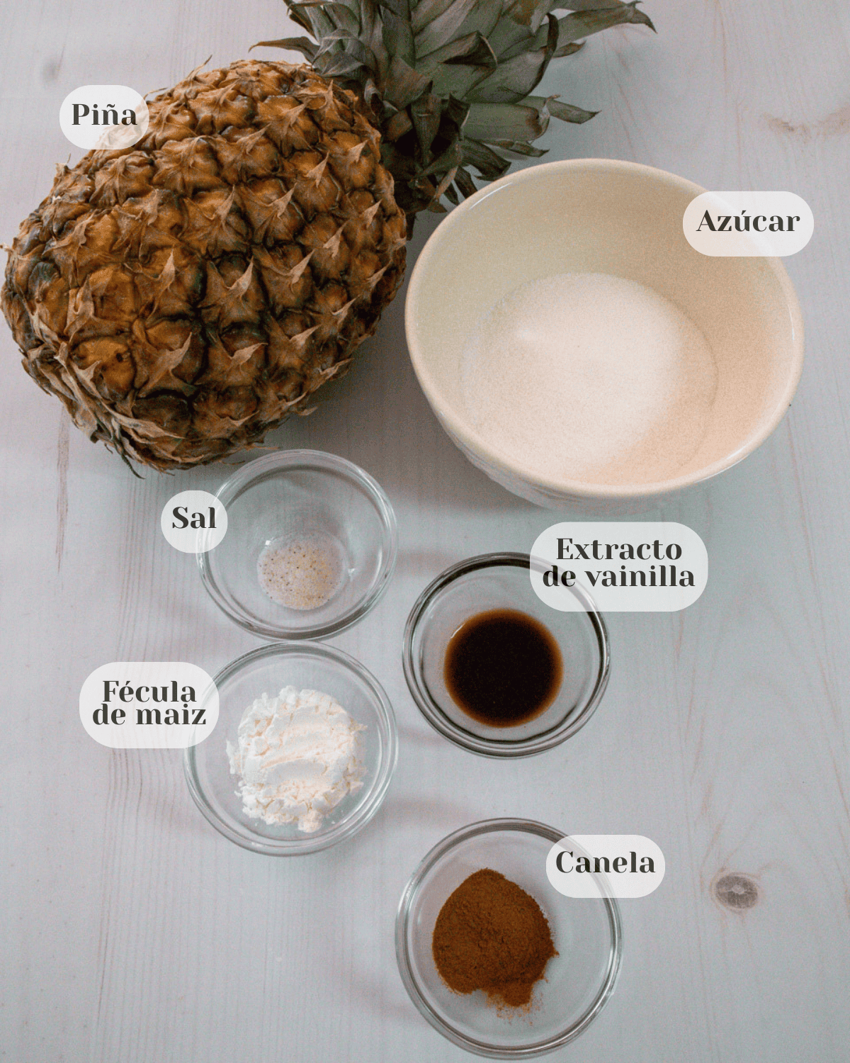 Se muestra una piña natural sin pelar, una taza de porcelana conteniendo azucar, y cuatro tacitas de vidrio transparentes conteniendo en casa una, sal, extracto de vainilla, fecula de maiz y canela.
