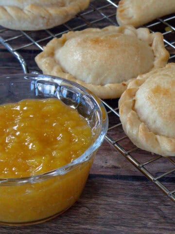 Se muestra una tacita de vidrio conteniendo jalea de piña, y en la parte trasera de la fotografia unas empanadas con firma circular.