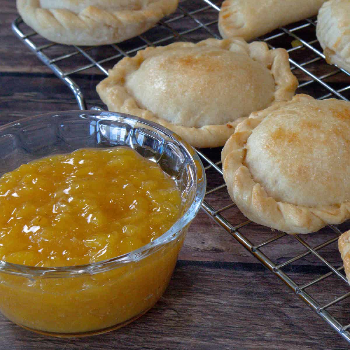 Se muestra una tacita de vidrio transparente conteniendo mermelada de piña. En la parte posterior de la fotografia se muestran unas empanadas en forma circular rellenas de jalea de piña.