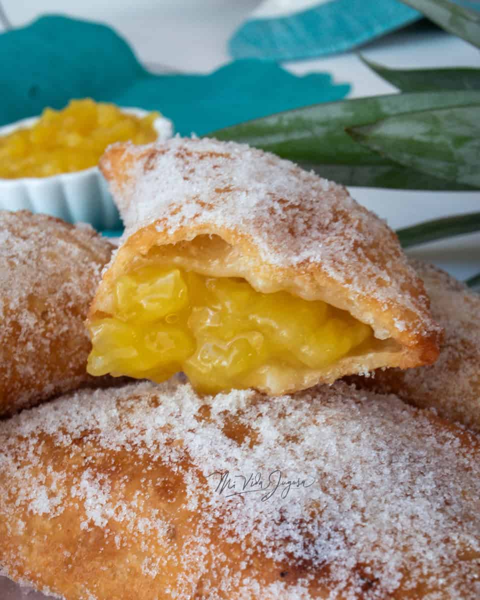 Fotografia de una pila de empanadas glaseadas de azucar resaltando en la parte de arriba una empanada partida a la mitad, desbordandose el relleno de piña.