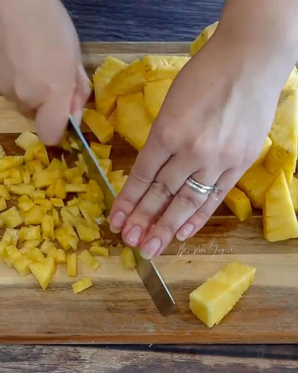 Fotografia donde se muestra una tabla de picar de madera con piña en cuadritos, unas manos sosteniendo el cuchillo mientras realiza los cuadritos.