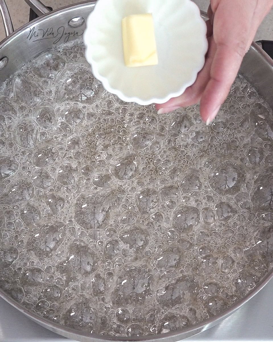 Un sarten conteniendo la mezcla de el agua y el azúcar de color ligeramente dorado, y una mano por encima sosteniendo una pequeña tacita con la mantequilla