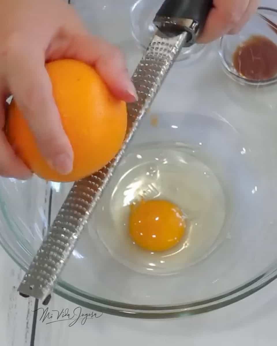 Un tazón de vidrio conteniendo un huevo, y en primer plano un séster rallando la cáscara de naranja 