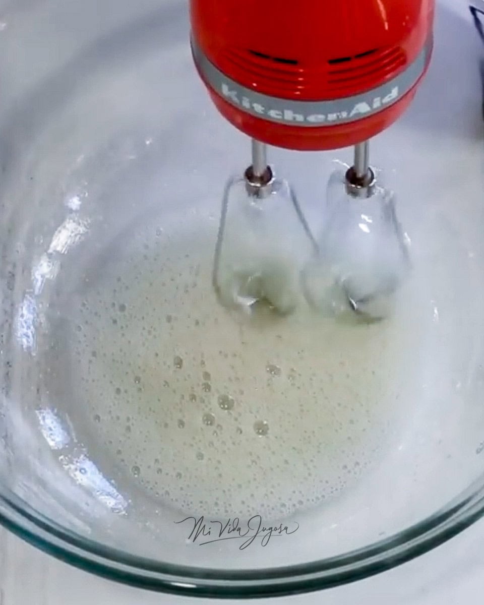 Un tazón de vidrio conteniendo la clara de huevo siendo mezclada por una batidora de mano