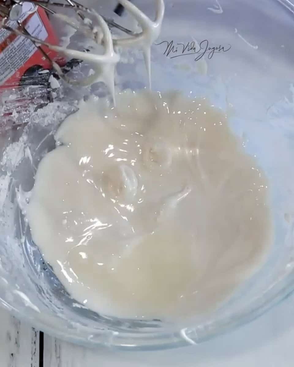Se muestra un bowl de vidrio conteniendo el glaseado listo, y las espatulas de la batidora de mano exchibiendo el punto ideal del glaseado.