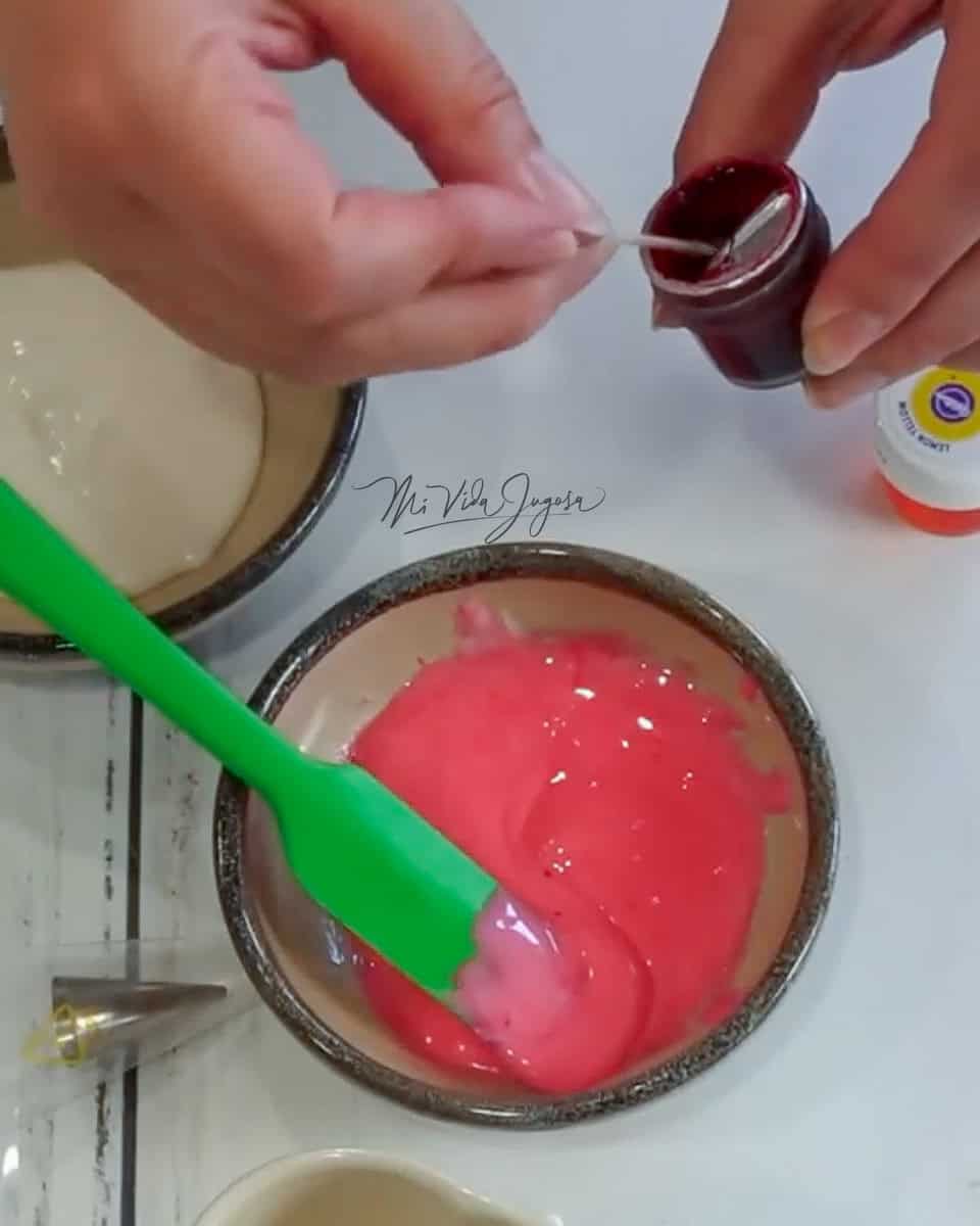 Un tacita con glaseado teñido de color rojo, y se ven las manos donde esta extrayendo colorante del tarrito de colorante