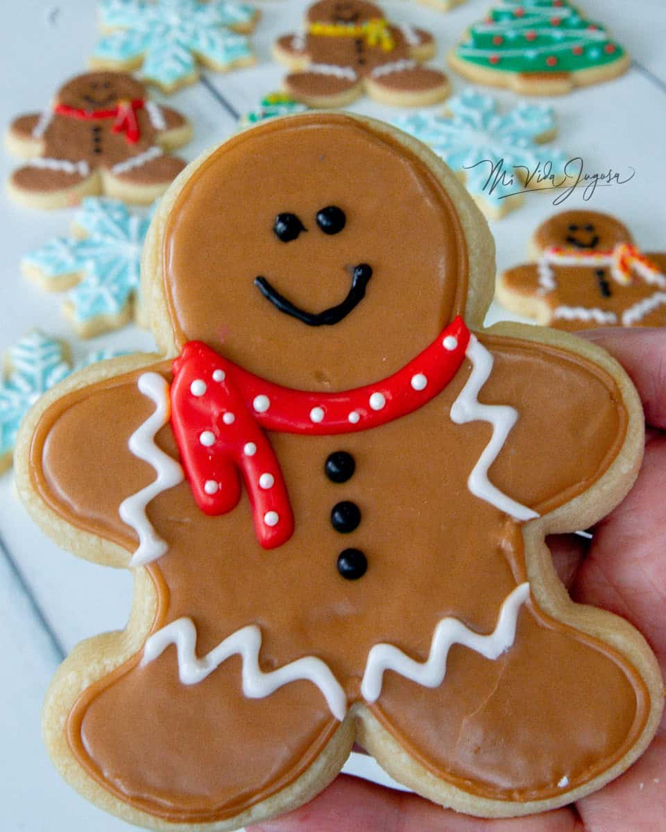 En primer plano una galleta de un muñeco de gengibre decorado con una bufanda, y por detras se muestran diferentes galletas navideñas