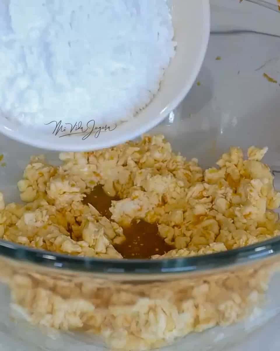 Un tazón de vidrio conteniendo la mezcla de huevo con mantequilla, y una taza agregando la azucar impalpable.