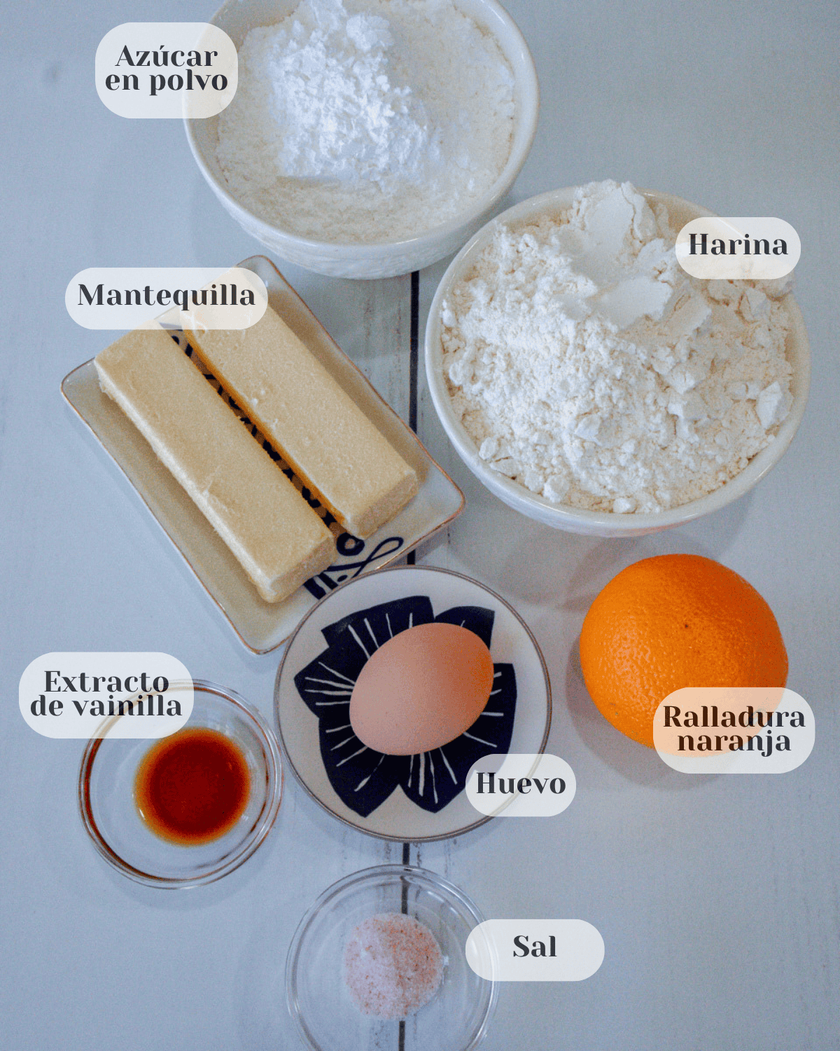 Se muestran diferentes recipientes pequenos conteniendo los ingredientes a utilizar: El azúcar, el harina, la mantequilla, la ralladura de naranja, el huevo, el extracto de vainilla y la sal 