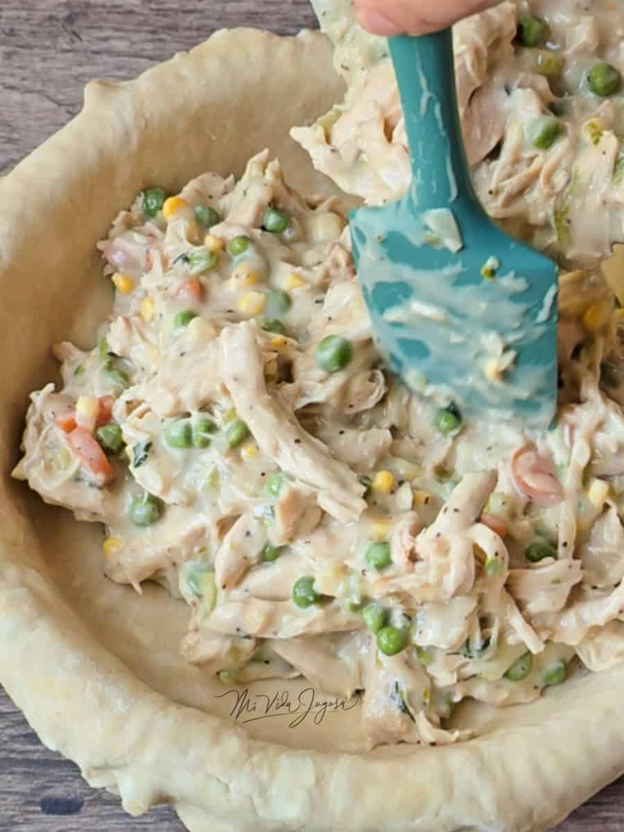 Un molde de pie sosteniendo la masa, una espatula acomodando el relleno de vegetales y pollo