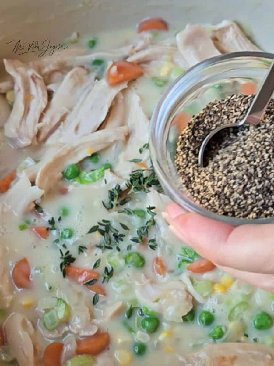 Mezcla del rellono de vegetales y pollo con unas hojas de tomillo al fondo de la imagen, mientas una mano sostiene una tacita que contiene pimienta negra y una cuchara de medida agarrando la pimienta