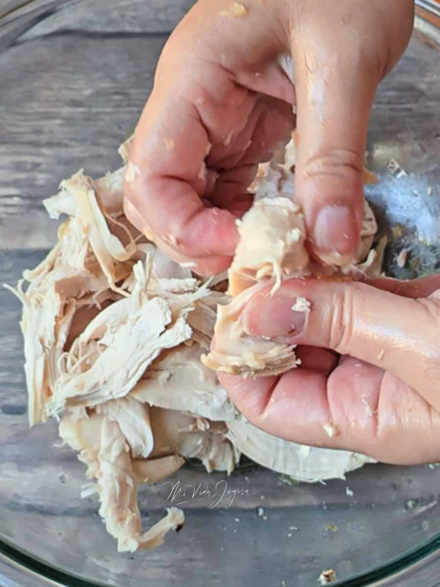 Dos manos desmenuzando un pollo que se encuentra en una tazon de vidrio
