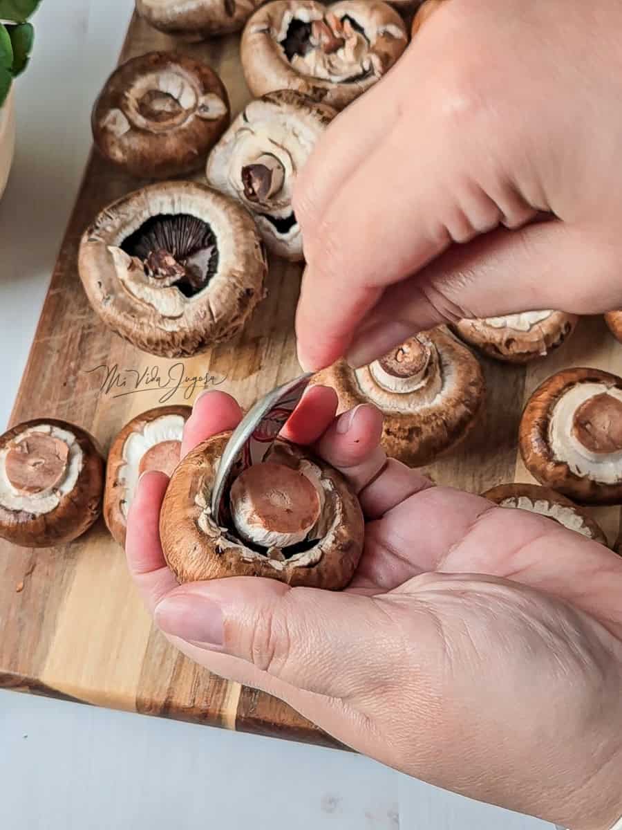 En primer plano una mano sostiene un hongo, con la otra mano se sostiene una cuchara que esta quitando el tallo al hongo, por detras se observa una tabla de picar con hongos