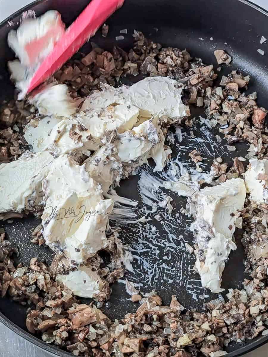 un sarten conteniendo el sofrito de la cebolla, los hongos y queso crema, siendo mesclados con una espatula
