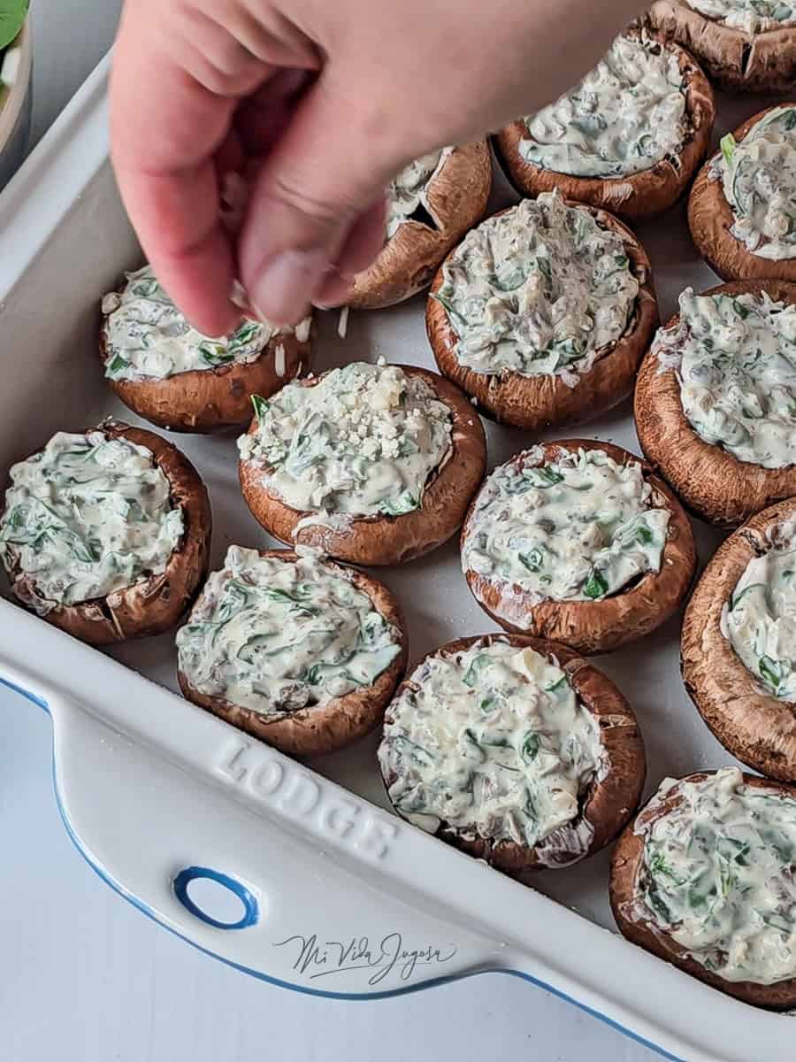 Un molde para hornear con hongos rellenos crudos, mientras una mano espolvorea queso parmesado rallado por encima