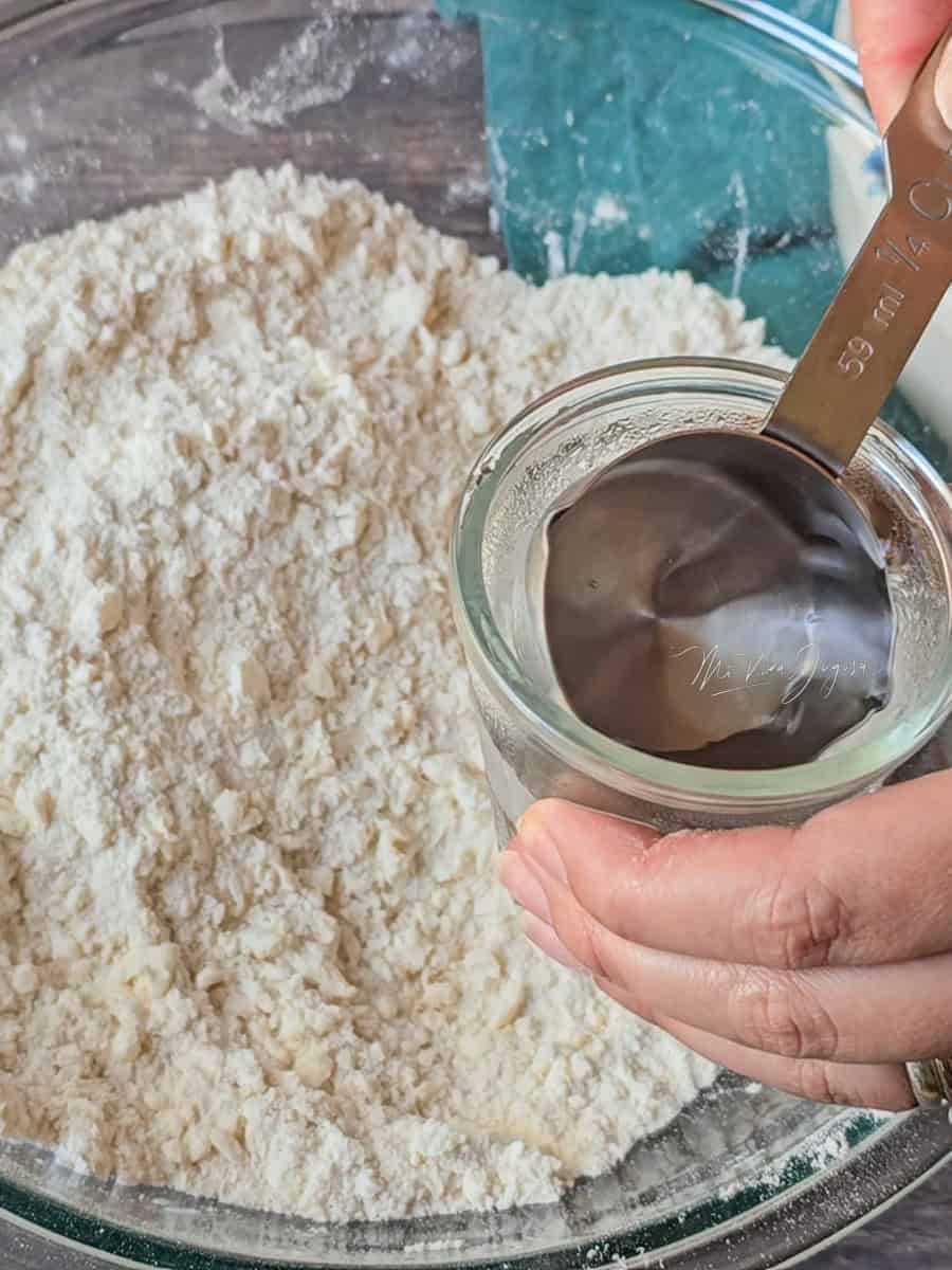 Un tazón con harina y trocitos de mantequilla, mientras una mano sostiene una tacita con agua fría y una taza medidora sumergida en ella.