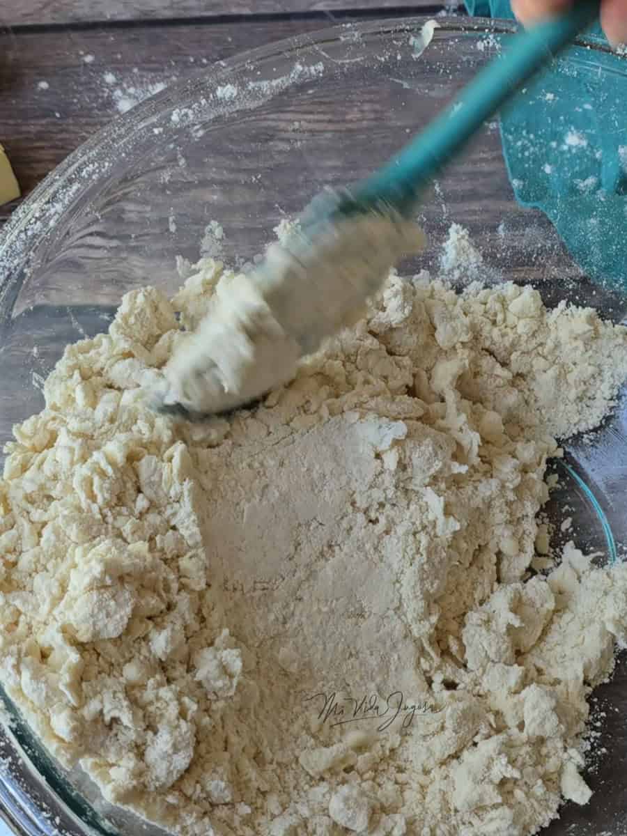 Un tazón con harina acompañado de una mano que sostiene una espátula lista para mezclar.