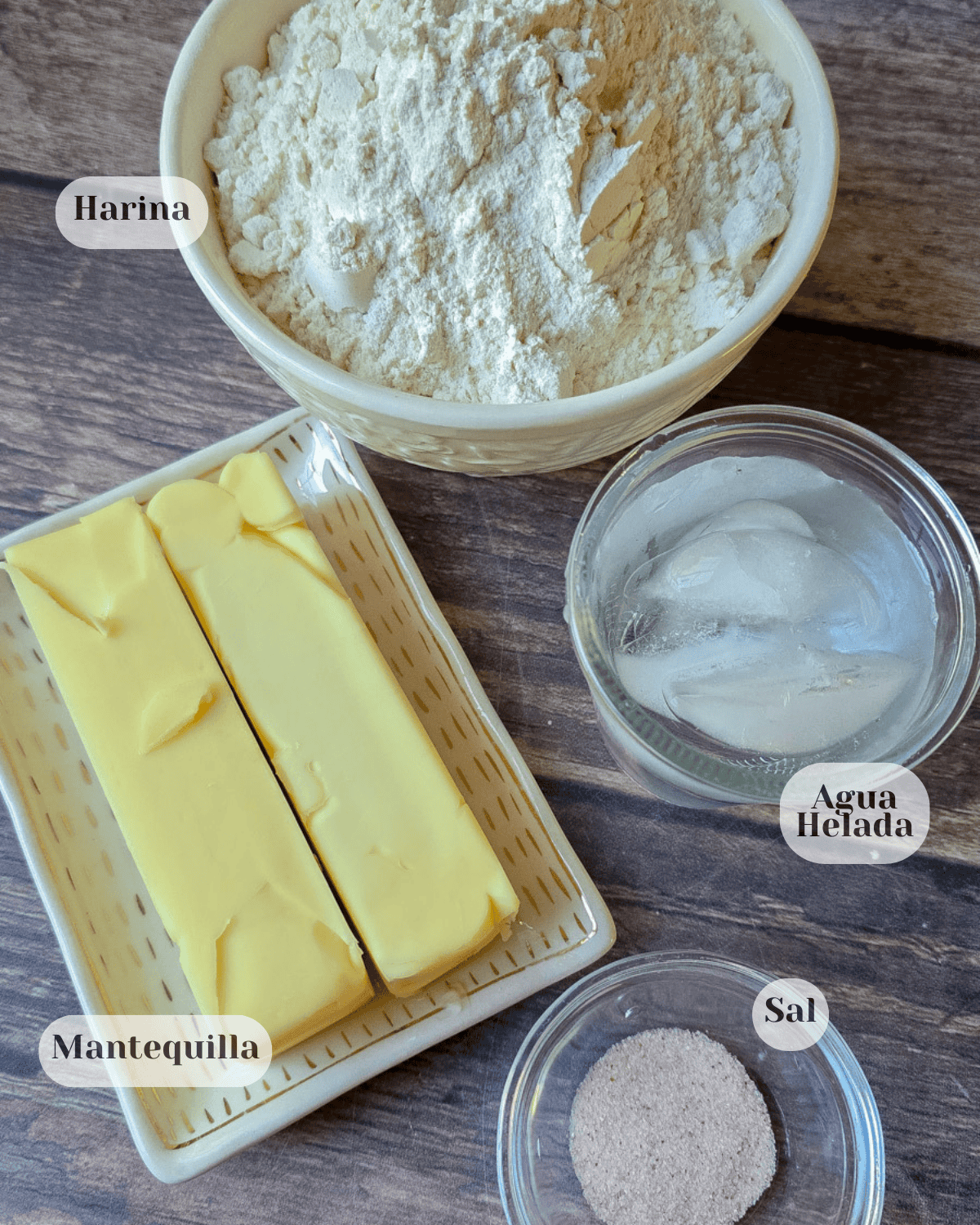 Una taza de harina junto a un plato pequeño con dos barras de mantequilla, una taza de agua con hielo y una pequeña tacita con sal.