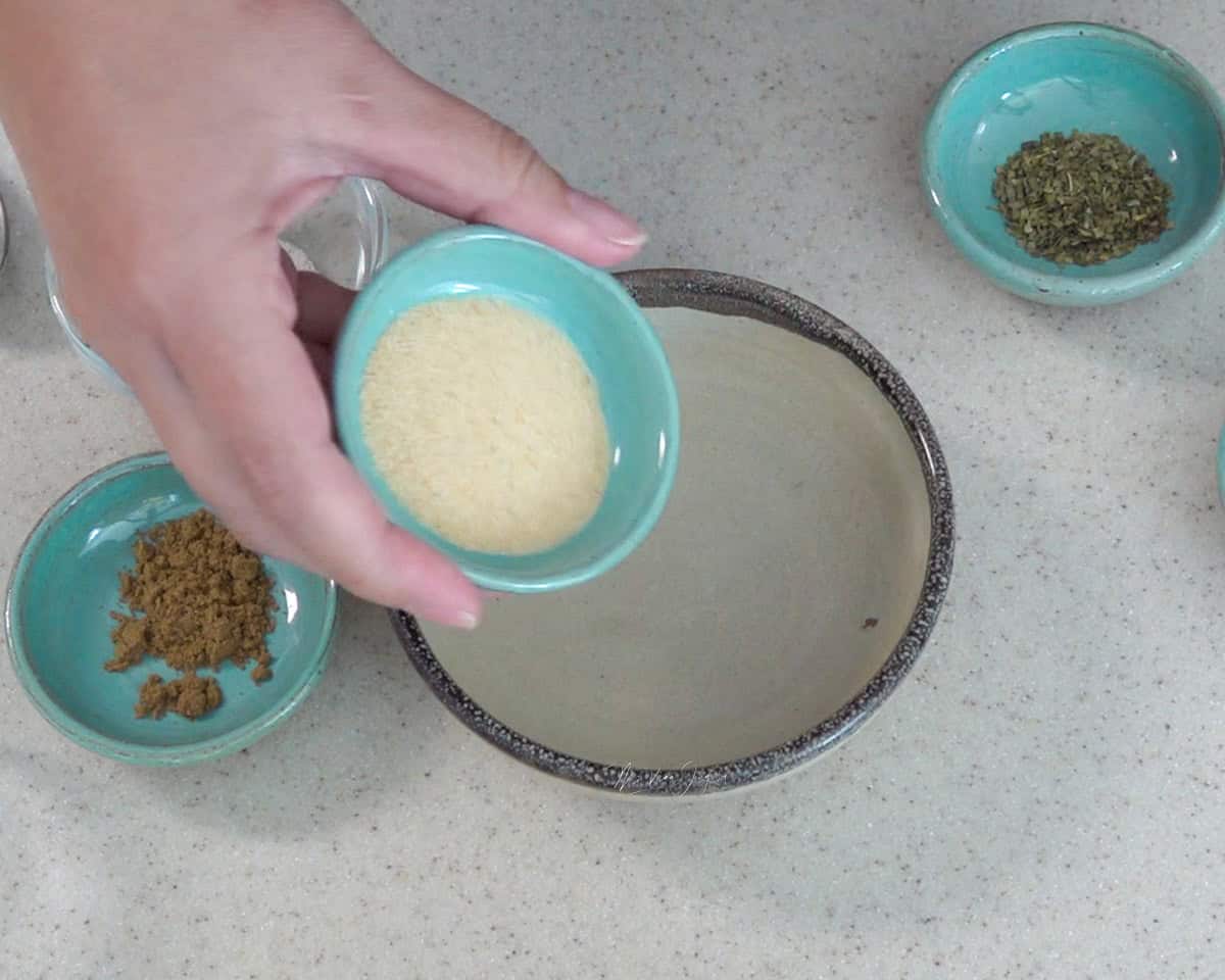 Una mano sostiene una tacita conteniendo ajo en polvo, por encima de una taza vacia, al lado dos tazas, una con comino y otra con orégano.