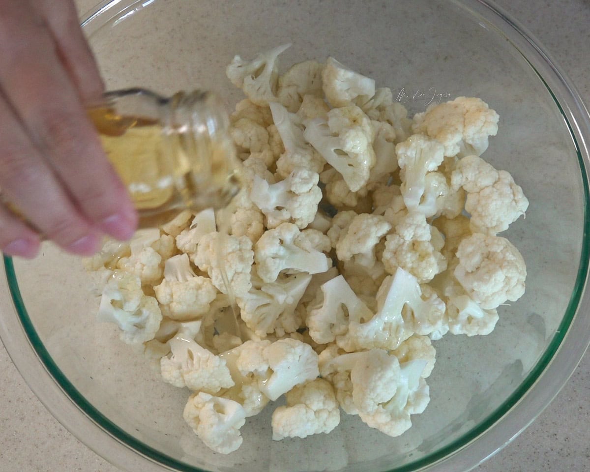 En un tazón con floretes de coliflor, una mano sostiene un vasito con aceite mientras lo vierte por encima.