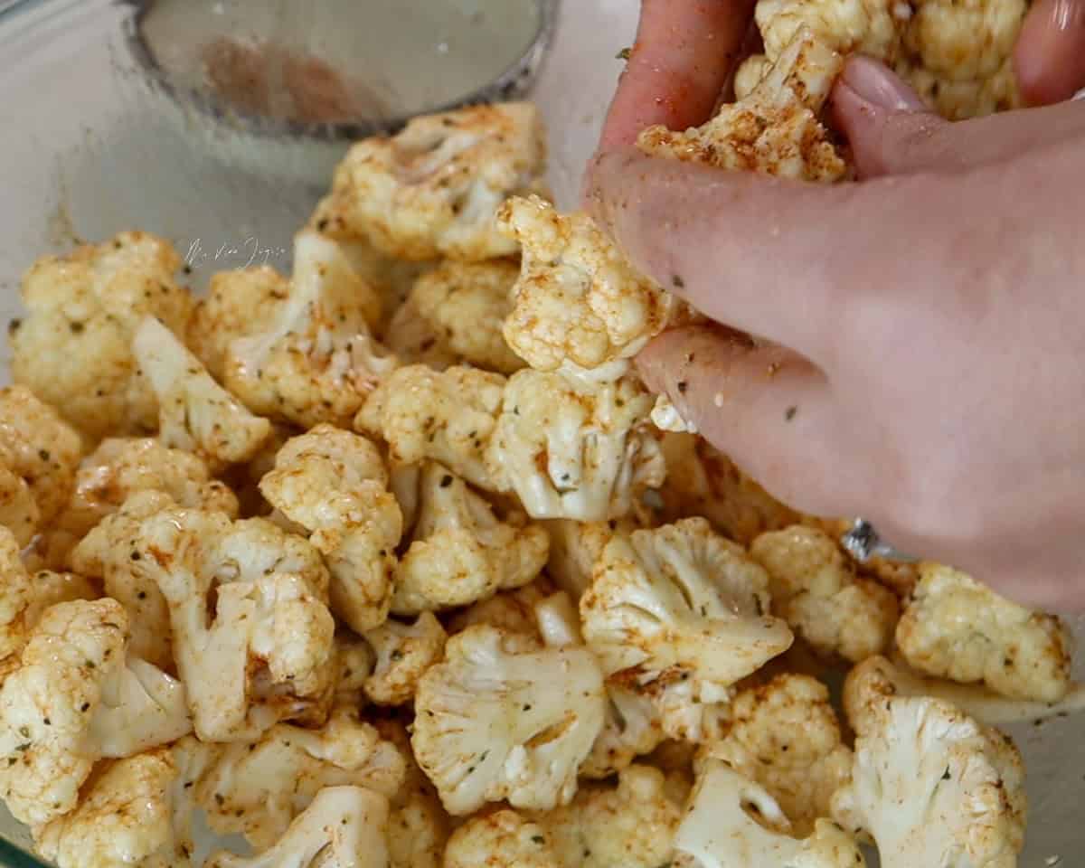 Unas manos mezclan y cubren con condimentos los floretes de coliflor.