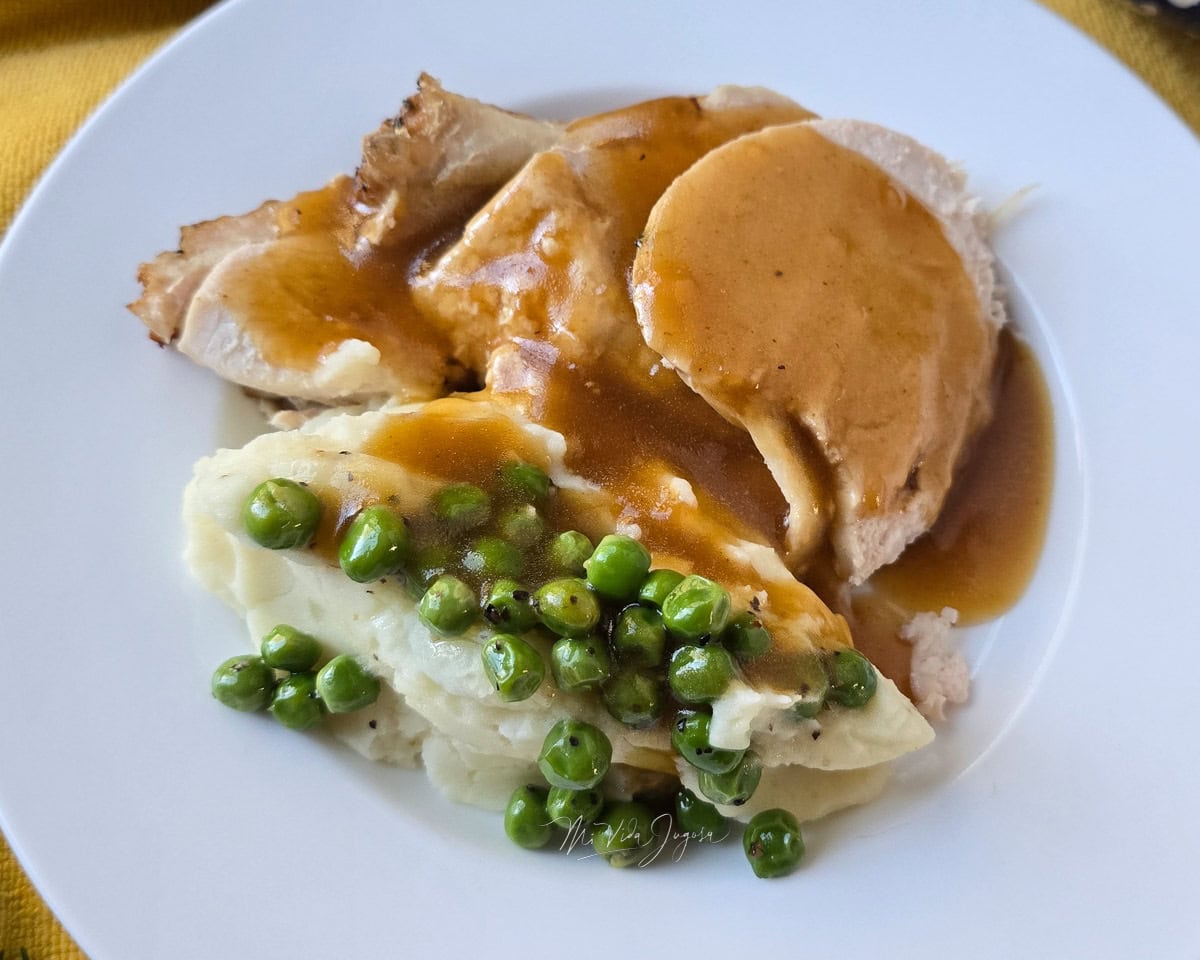 Un plato servido con lonjas de pavo bañados en gravy, acompañado de pure con arvejas encima, por detras de la fotografia un platon que contiene la pechuga de pavo